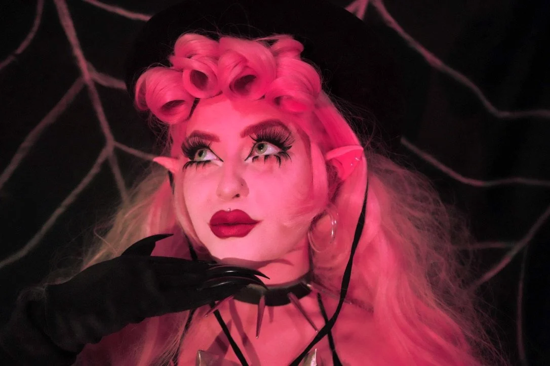 A woman with pink hair styled in retro curls, wearing theatrical makeup with exaggerated eye makeup and dark lipstick, and black gloves. She has elf-like ears and large hoop earrings, sitting against a dark background with web-like patterns.