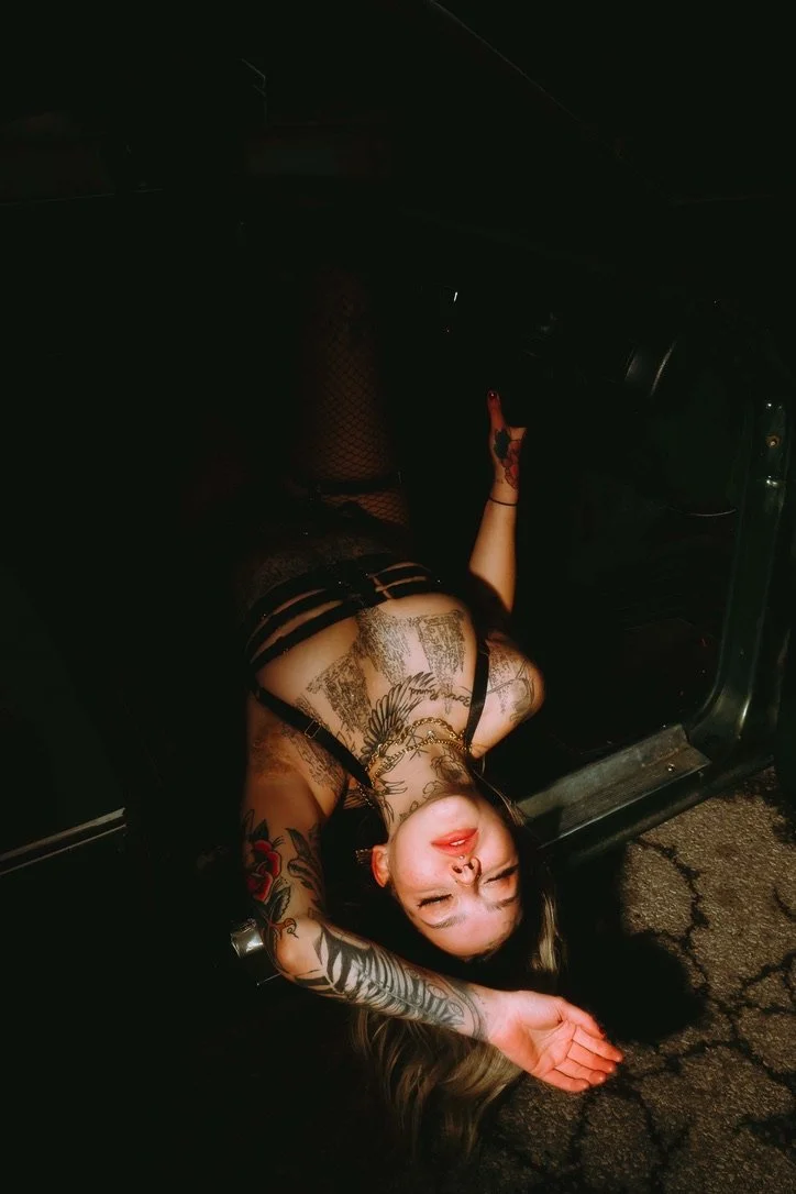 A woman with tattoos, wearing fishnet stockings and black lingerie, lying on the ground underneath a car, making a face with one eye closed and her tongue out.