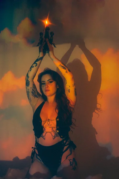 A woman with tattoos is holding a lit flare above her head, creating a shadow on the wall behind her.