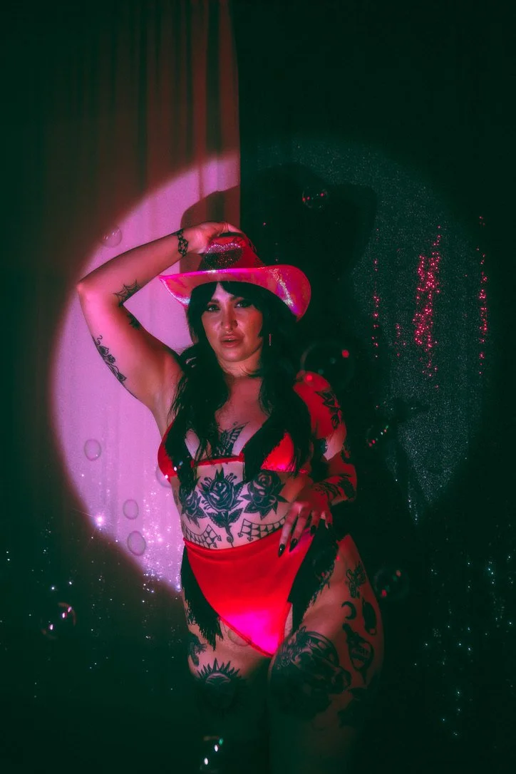 Woman wearing a red lingerie set and a pink cowboy hat, with multiple tattoos, standing against a dark background with pink lighting and sparkles.