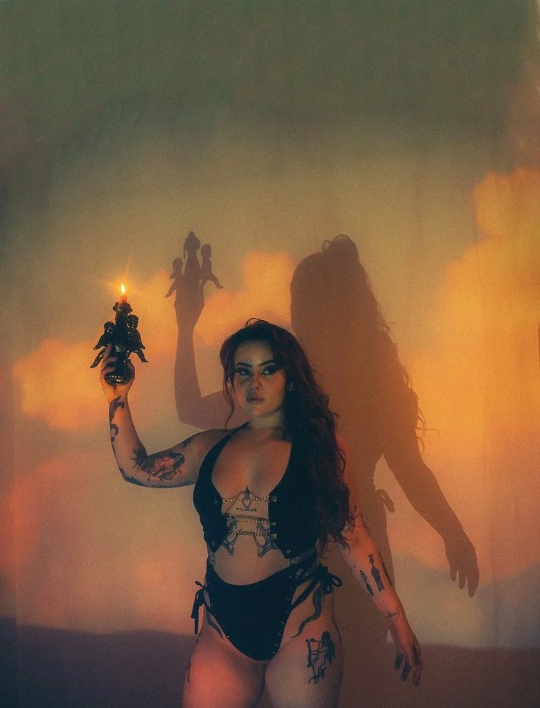 A woman with long dark hair and tattoos holding a lit candle, standing against a warm orange background with her shadow cast on the wall.