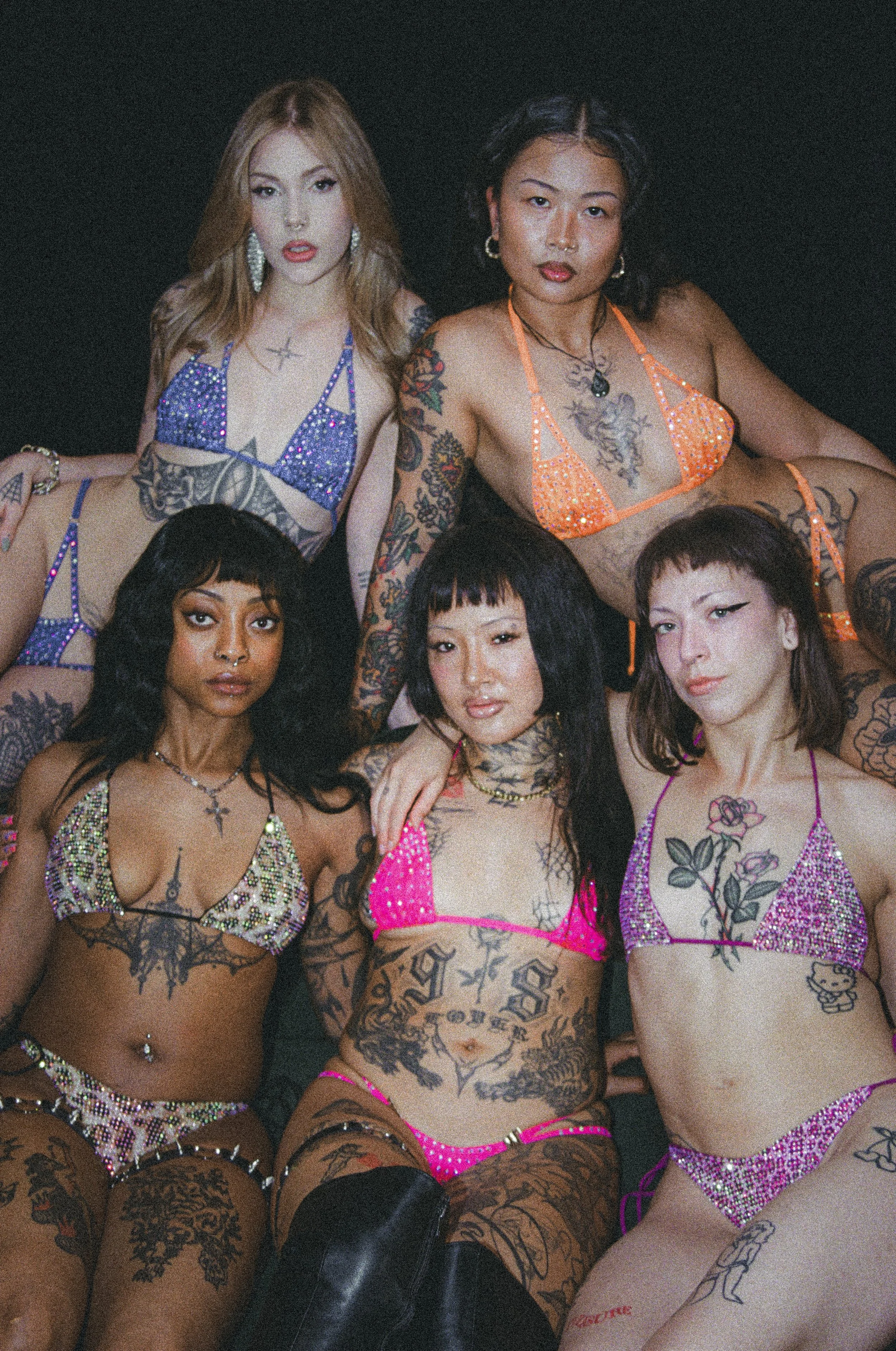 Four tattooed women pose together in colorful rhinestone bikini tops against a dark background.