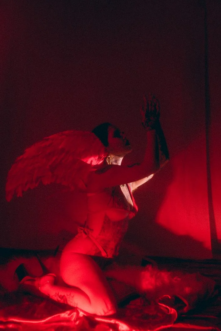 A person with angel wings kneels on the floor, illuminated by red lighting, with one arm raised and head tilted back, in a dimly lit room.