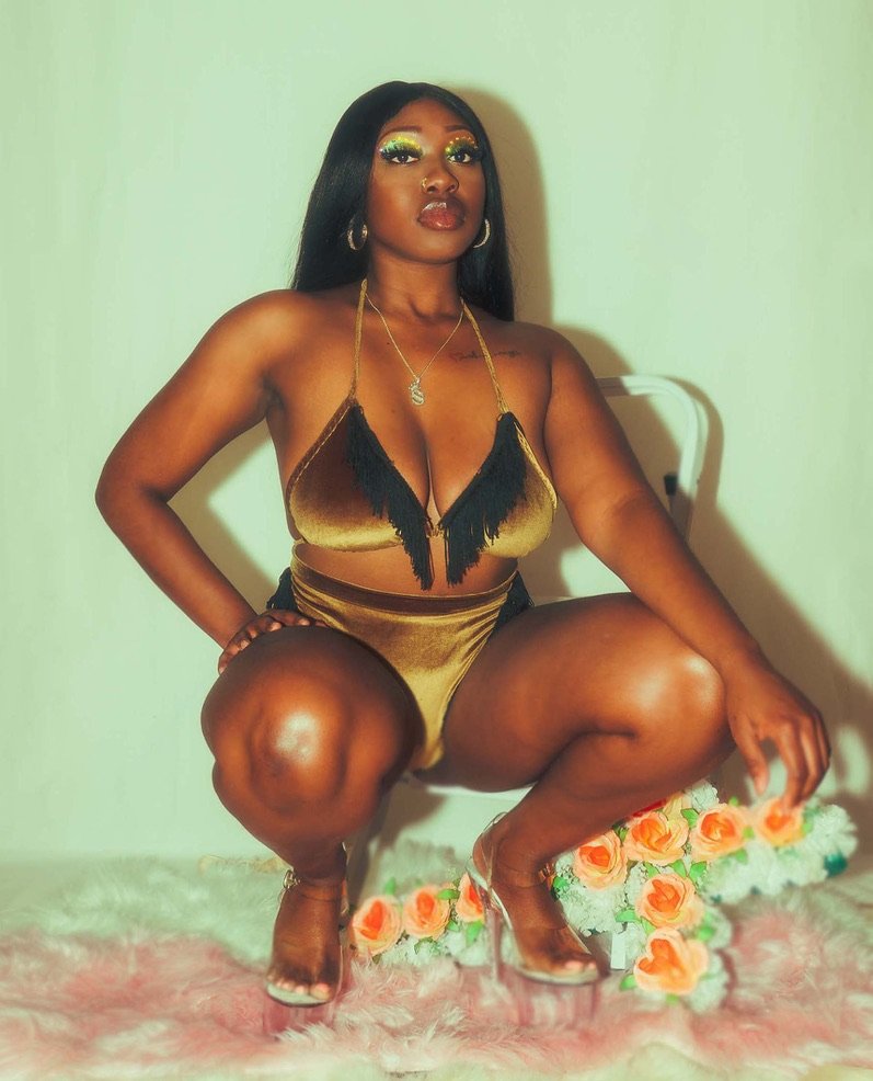 A woman masked in golden velvet outfit with black fringe, squatting on a decorated floral carpet, with makeup, jewelry, and high heels, posing against a plain wall.