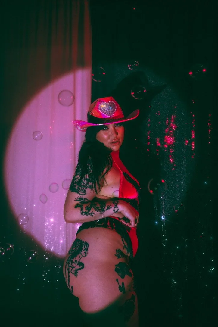 Woman with dark hair wearing a pink cowboy hat with a heart design, red and black lingerie, and tattoos on her arm and thigh, standing against a dark background with pink and green lighting effects and bubbles.