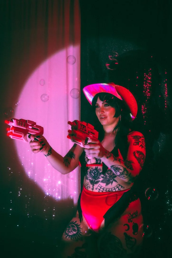 A woman with tattoos on her arms, legs, and torso, wearing a pink cowboy hat and red lingerie, holding two toy guns. She is standing in front of a colorful background with pink and purple lighting.