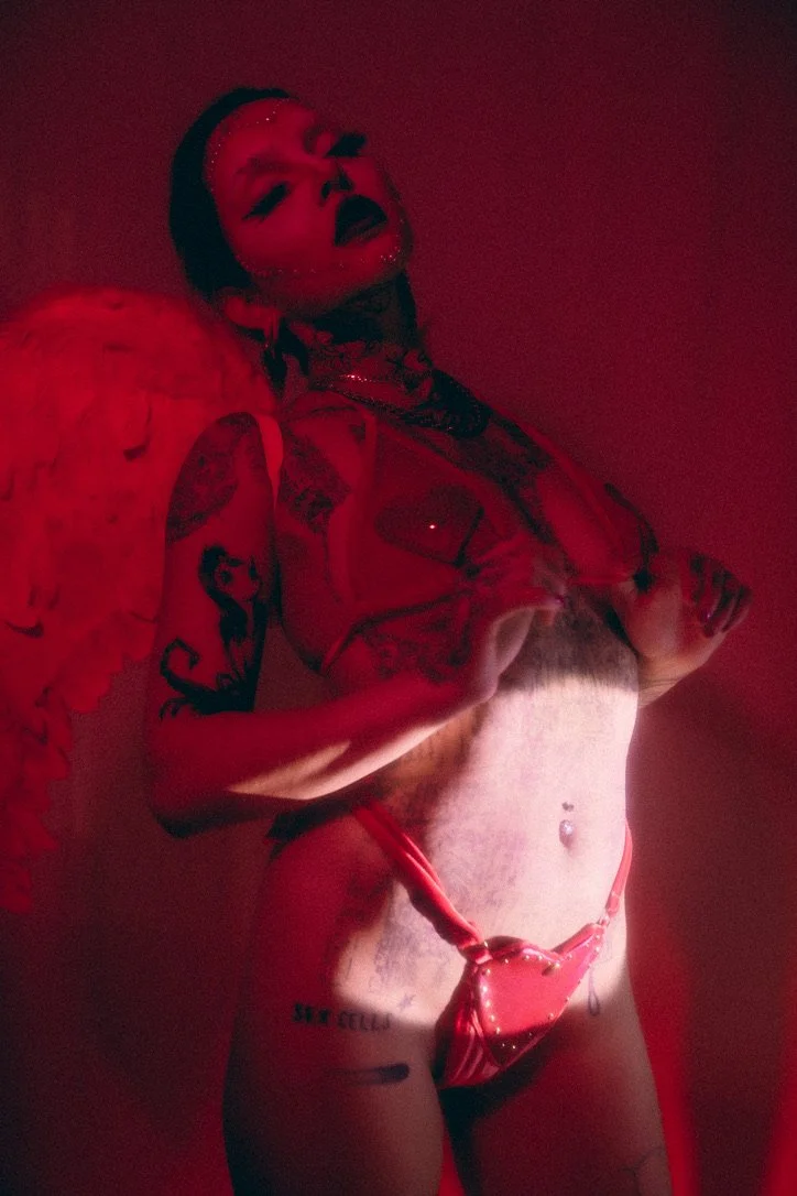 A person with tattoos, piercings, and makeup posing in red lighting, wearing red vinyl lingerie, with an angel wing accessory on their back.