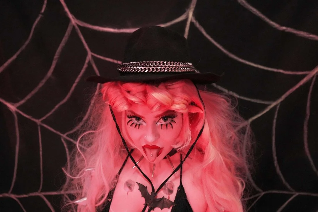 A woman with pink hair styled in loose waves, wearing dark eye makeup and bright red lipstick, sticking her tongue out. She is dressed in a black outfit with a spider necklace, wearing a black hat decorated with chains, and standing in front of a spi