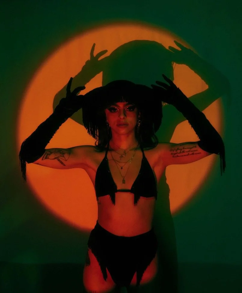 Woman in black lingerie with tattoos, wearing long black gloves, holding a wide-brimmed hat, casting a shadow against orange and green lighting.