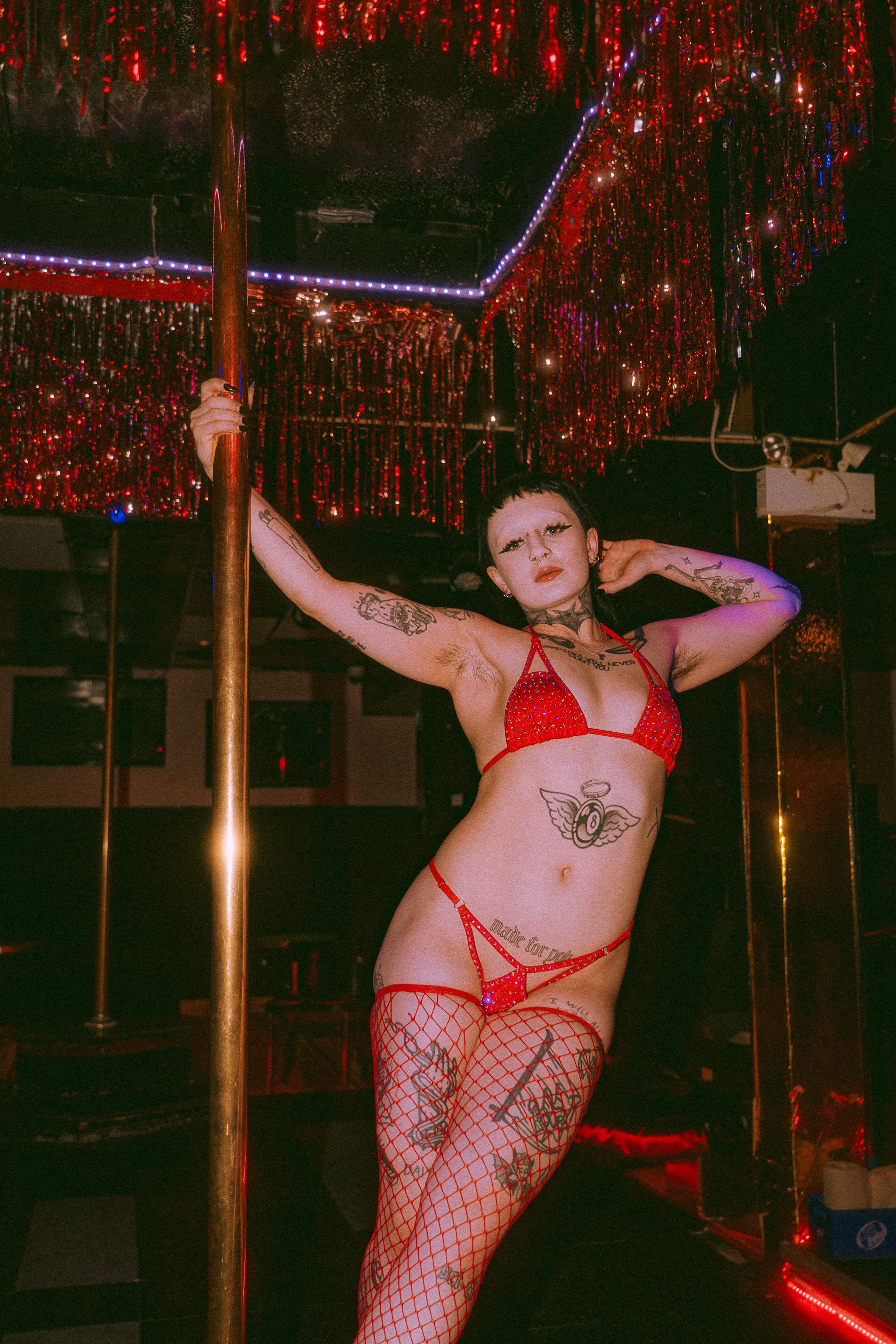 A person in a red showing outfit with tattoos on their body, standing next to a golden pole in a dimly lit room with red and purple lighting, and red and silver shiny decorations overhead.