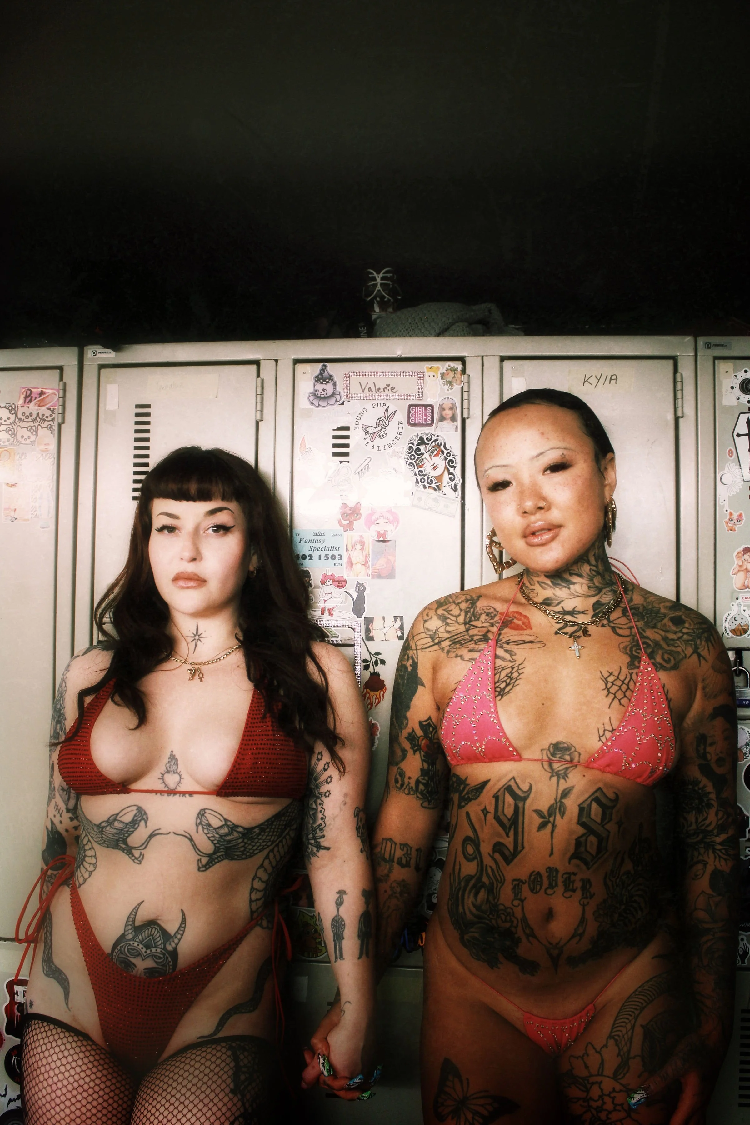 Two women with tattoos and revealing clothing standing in front of a row of lockers decorated with stickers and drawings, in a dimly lit setting.