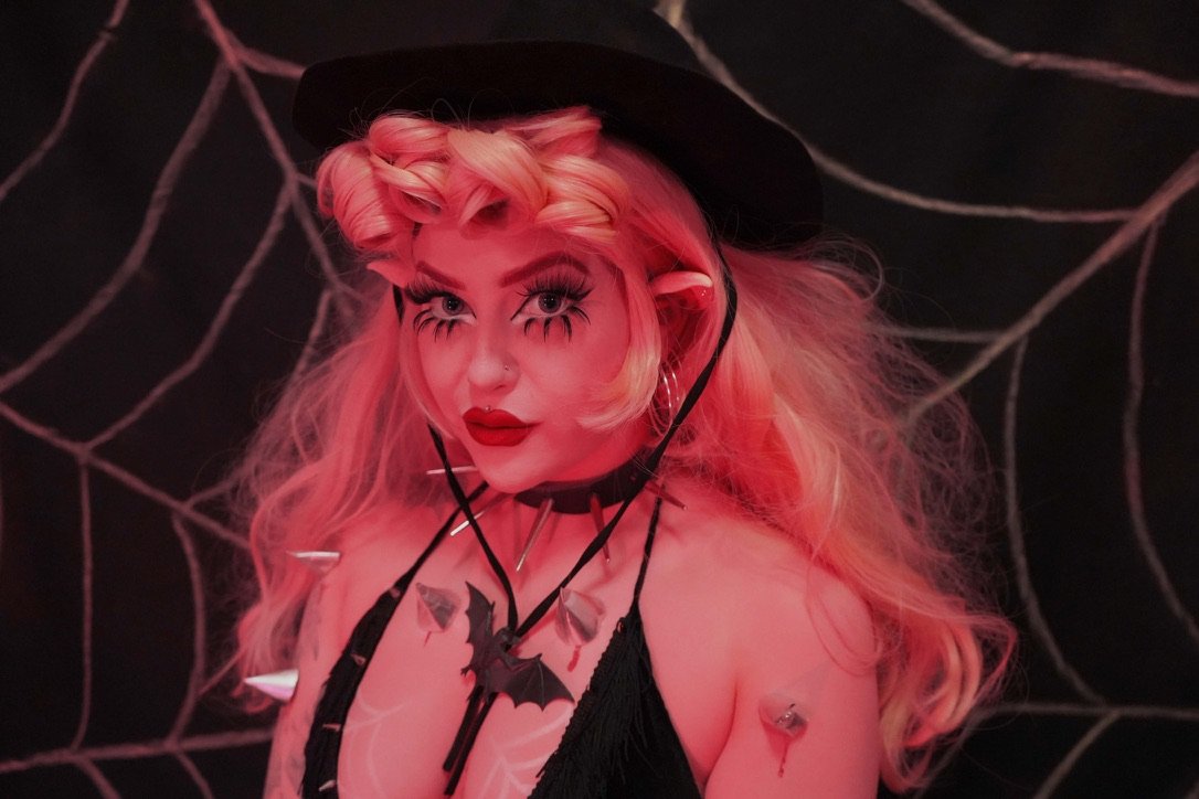 Person in Halloween costume with spider web background, wearing a witch hat, with dramatic makeup and accessories.