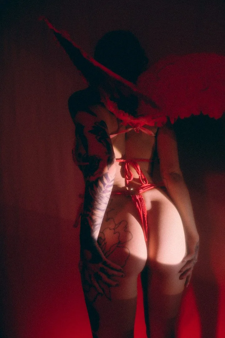 A person dressed as a devil or angel with large red wings, wearing red lingerie and a black hat, standing with their back to the camera in a dimly lit room with red lighting.