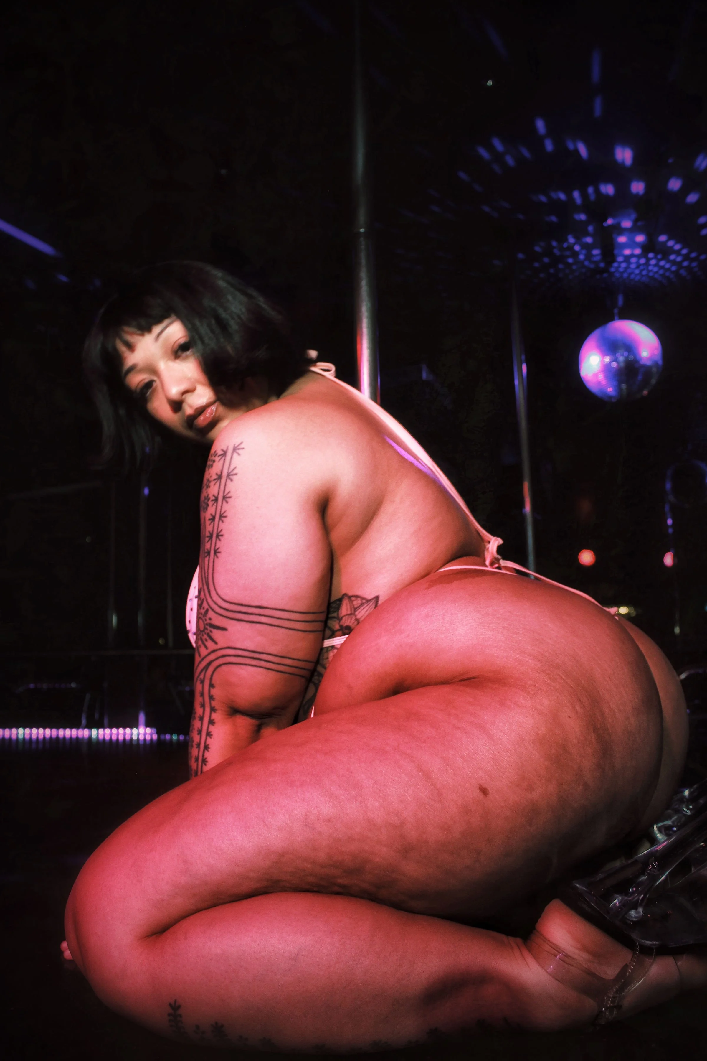 A woman with black hair and tattoos poses in a nightclub with purple lighting and a disco ball overhead.