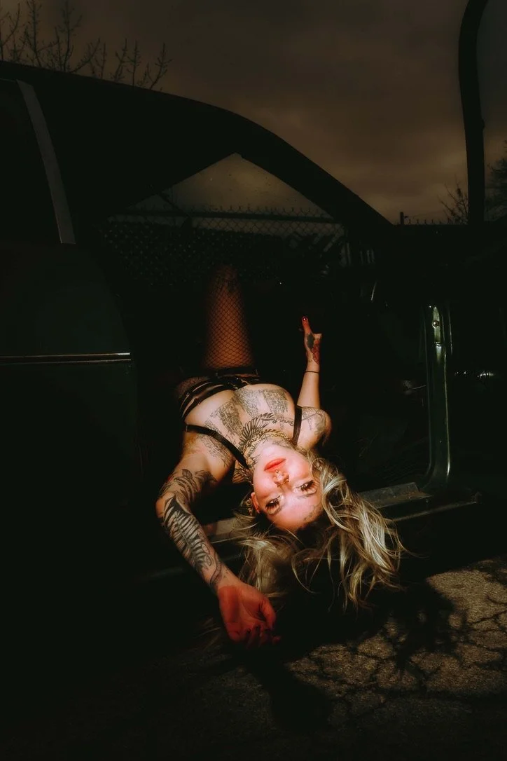 A woman with tattoos lying upside down on the floor outside a vehicle at night, with her head near the vehicle's open door, looking at the camera.