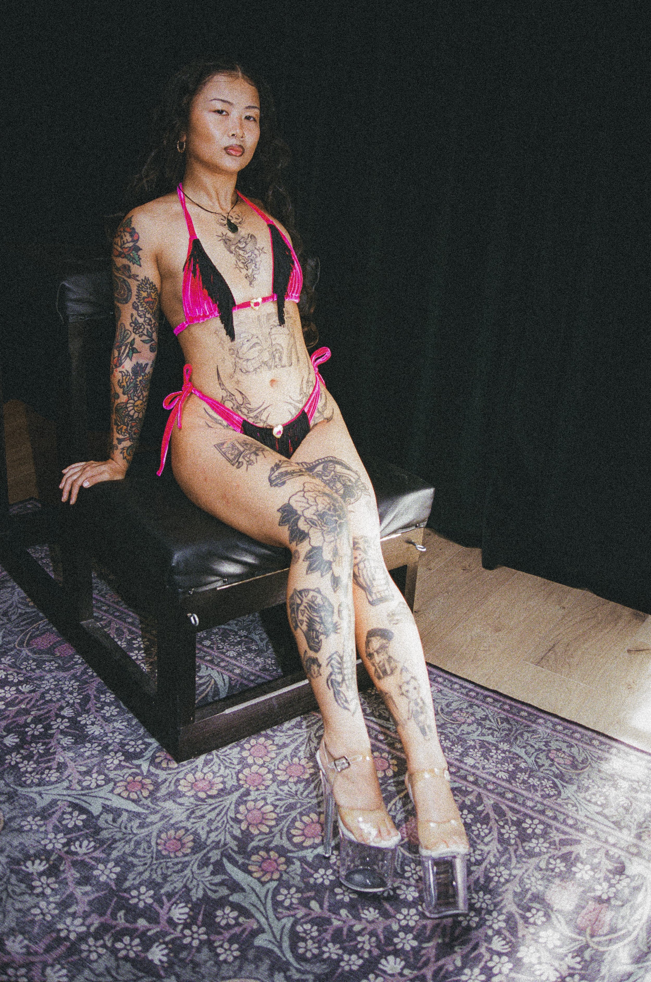 Tattooed woman in a black and hot-pink lingerie outfit posing on a black couch in a dimly lit room.