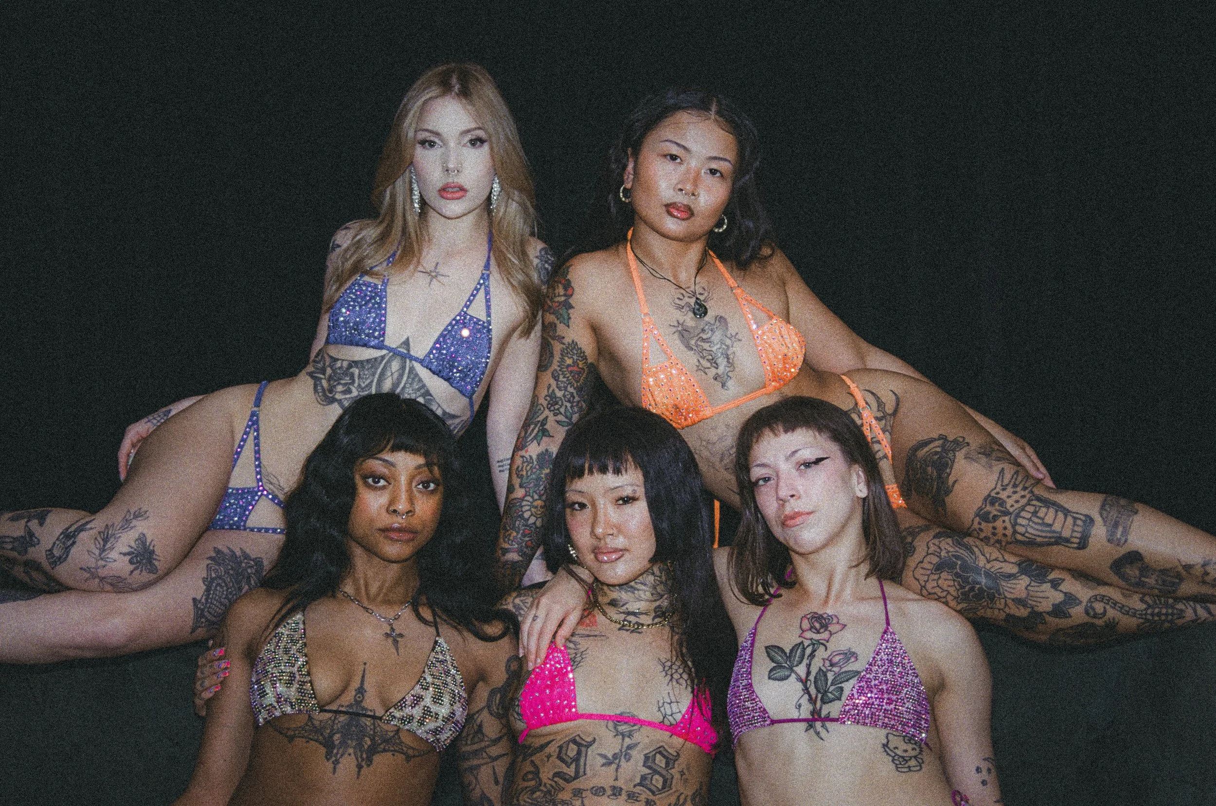 Five tattooed women pose together in colorful rhinestone bikinis against a dark background.
