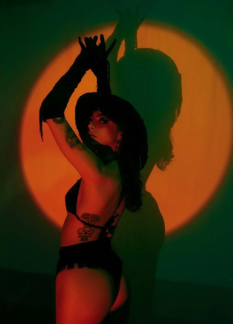 A woman with dark hair, tattoos, and earrings poses in lingerie with one arm raised. Her shadow is cast on the wall behind her, against a green and orange background.