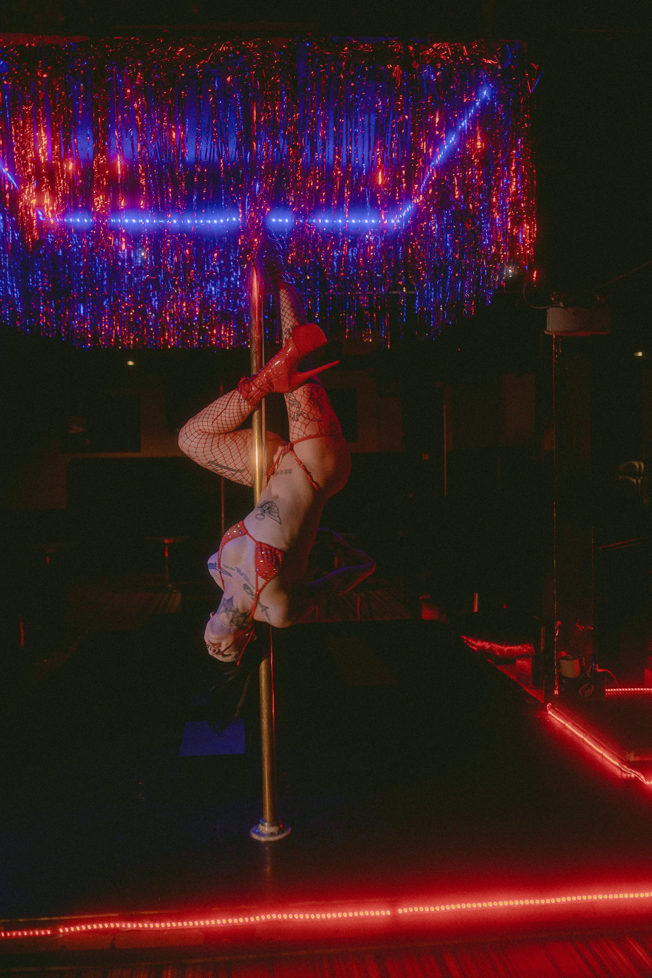 A pole dancer performs on a vertical pole in a dimly lit club with neon red and blue lighting. She is wearing fishnet stockings, red high-heeled shoes, and revealing outfit, with her head upside down and legs wrapped around the pole.