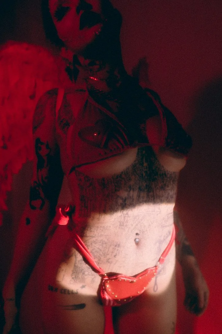 A woman taking a mirror selfie in a dimly lit room with red lighting. She has tattoos on her arms, chest, and stomach, and is wearing a black lace top, red thong, and a small red bag hanging from her waist.