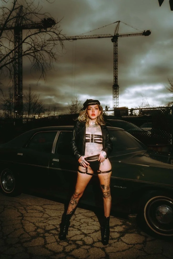 A young woman with curly blonde hair posing in front of a vintage black car during a cloudy, overcast day. She is wearing a black leather jacket, fishnet stockings, black boots, and a black cap, with a cityscape and construction cranes in the backgro