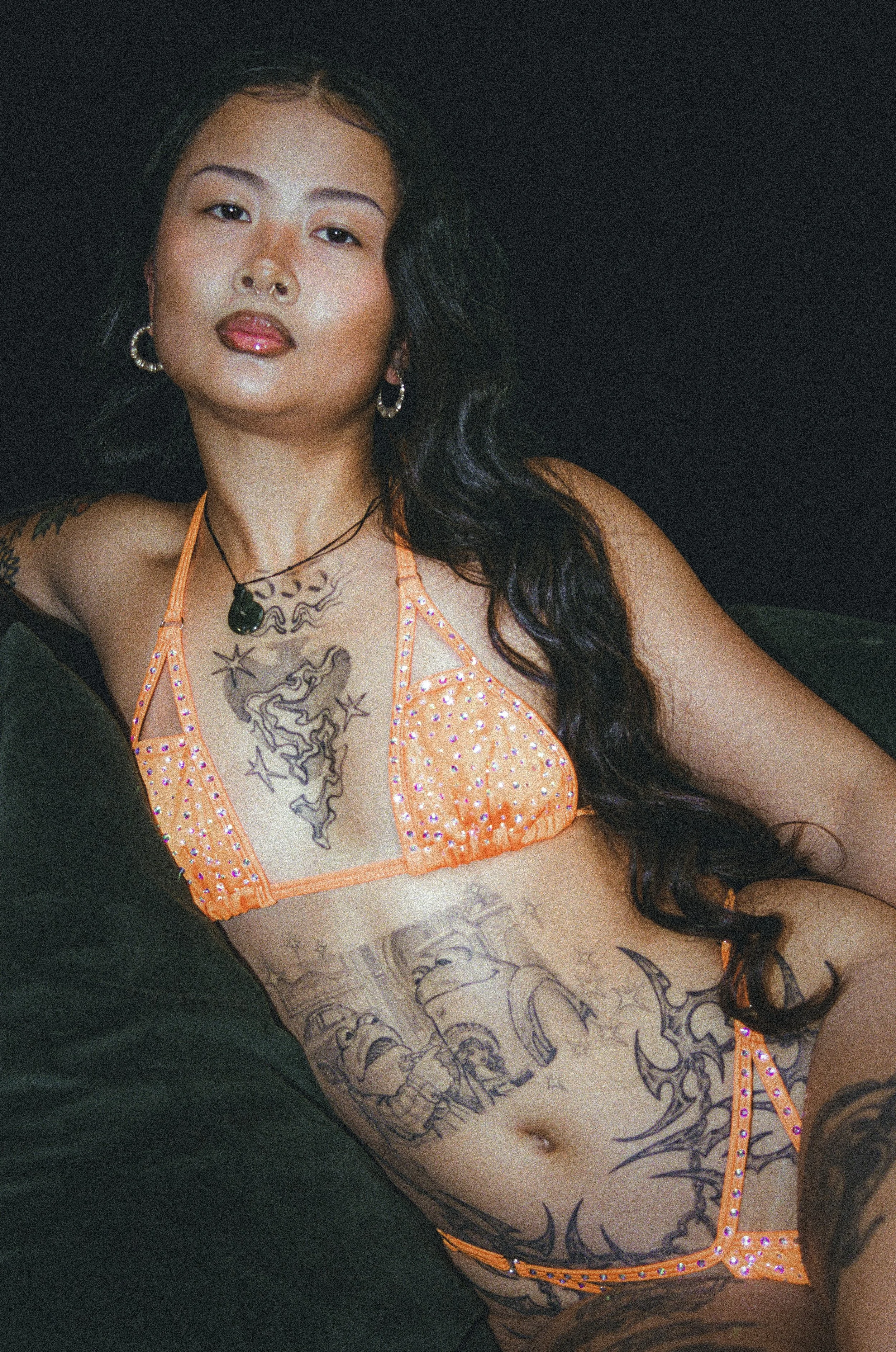 Close-up portrait of a woman with long dark hair, hoop earrings, tattoos, and a rhinestone-studded orange top against a dark background.