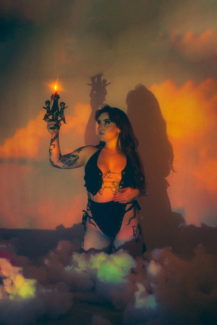 A woman with tattoos in black lingerie and a vest holding a lit candle, surrounded by colorful clouds or smoke, casting a shadow on the wall behind her.