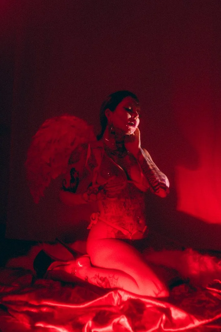 A woman with tattoos and dark hair poses on a bed in red lighting, wearing a corset, thigh-high stockings, and high heels, with a large feathered accessory on her back.