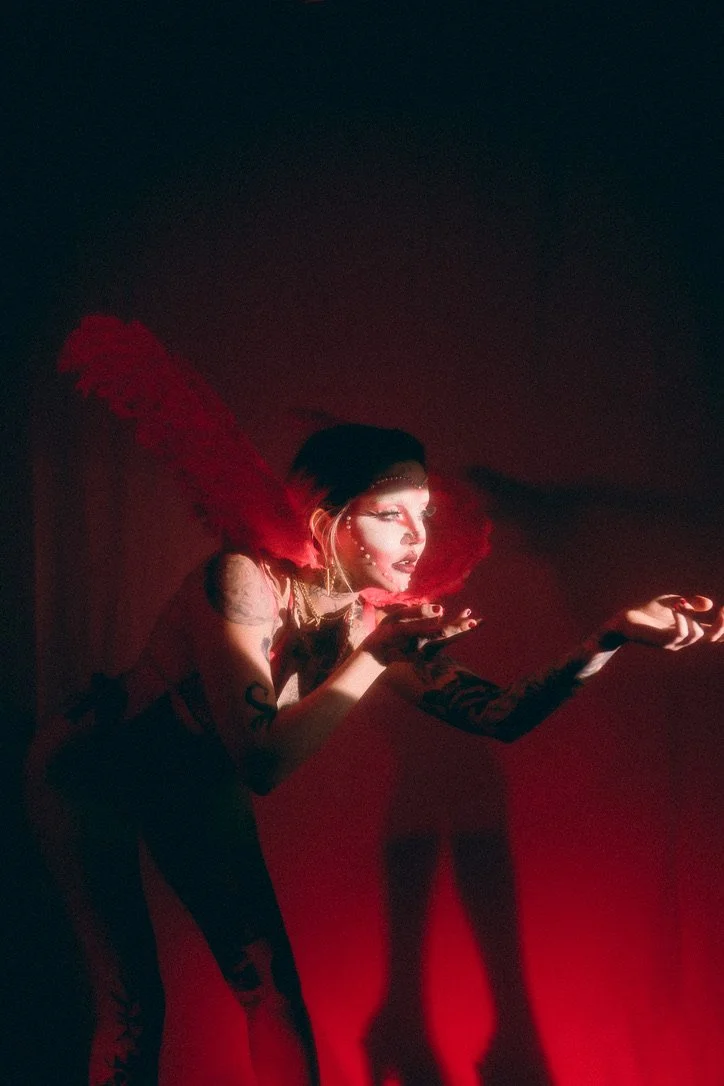 A woman with dramatic makeup and tattoos, wearing a black headpiece with a large red feather, is blowing a kiss. She is dressed in black with fishnet stockings and has long black hair. The background is dark with red lighting.