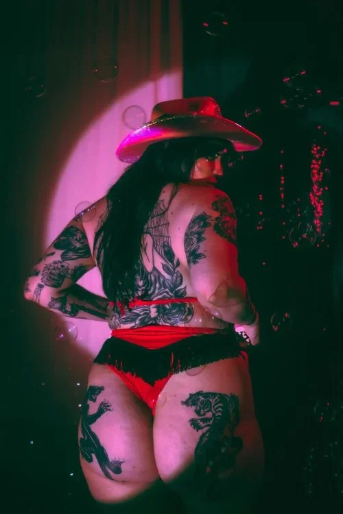 A woman with tattoos wearing a cowboy hat, red fringe shorts, and red underwear, standing with her back turned in a dimly lit room.