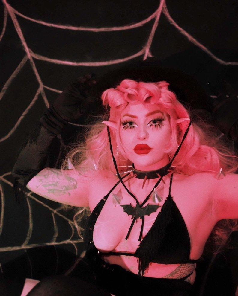 A woman dressed in a Halloween costume with pink hair, elf ears, and vampire makeup, surrounded by spider webs.