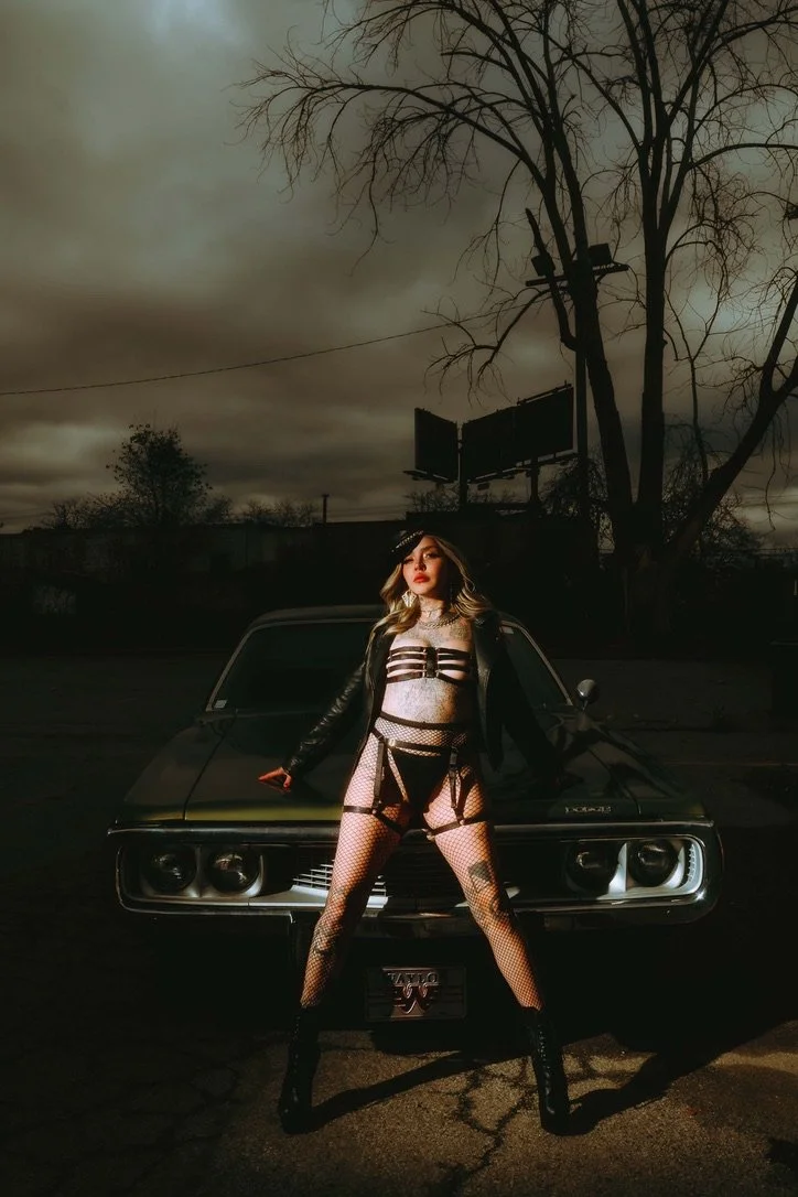Woman in edgy black leather and fishnet outfit standing with legs apart in front of a classic black muscle car during a cloudy, dark evening with a leafless tree and billboard in the background.