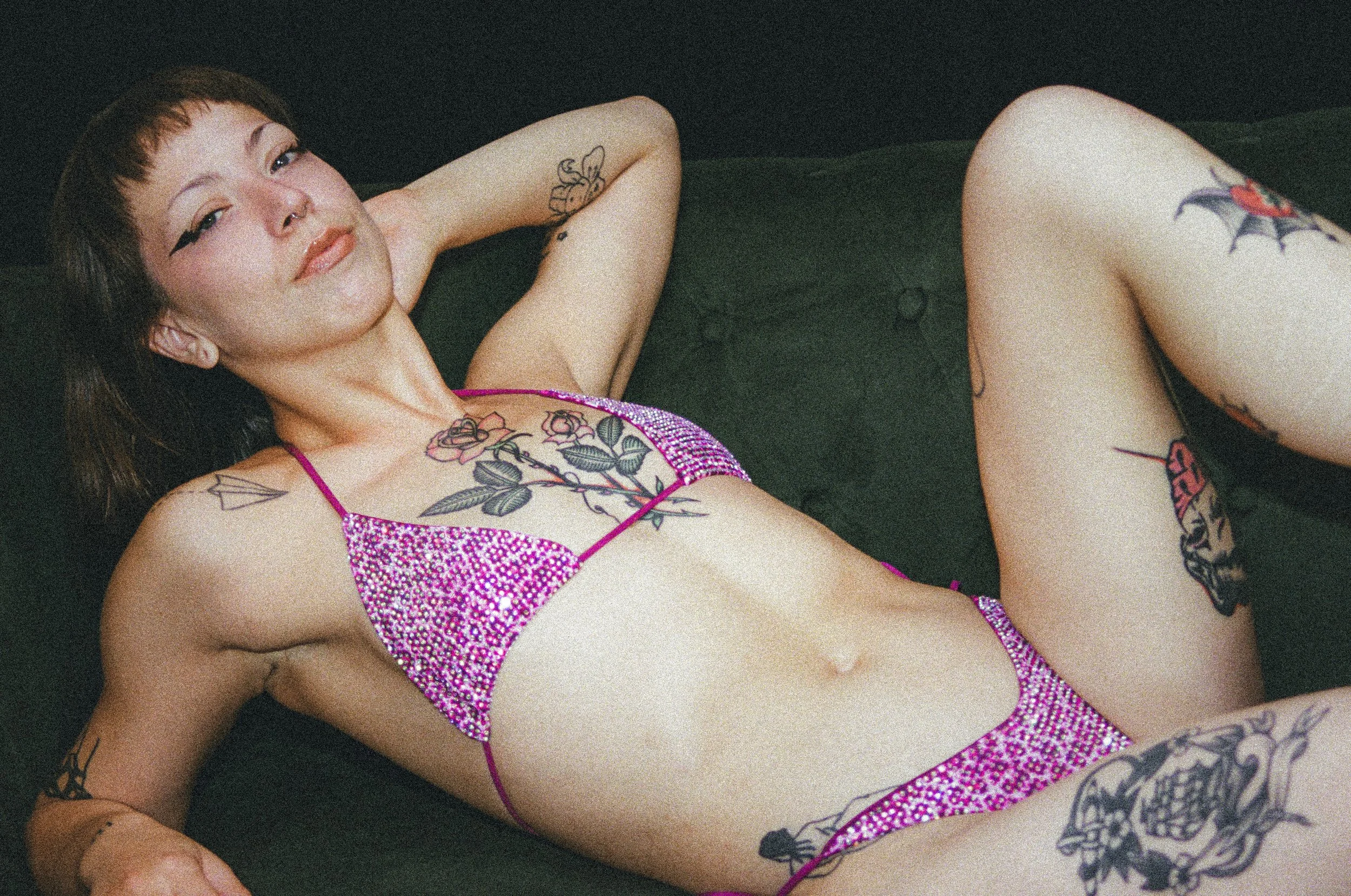 Tattooed woman reclining on a dark surface in a sparkling pink bikini, looking at the camera.