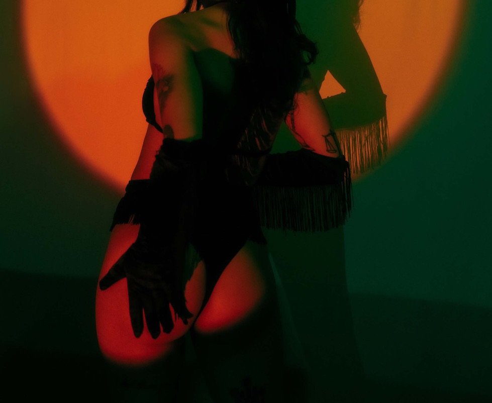 A woman with long dark hair posed against a colorful, projected background with orange, green, and teal hues, wearing black gloves and an outfit with fringe details.