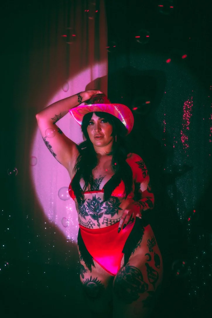 Woman with long dark hair and multiple tattoos, wearing red lingerie and a matching wide-brimmed pink hat, posing in front of a dark background with floating bubbles and red light reflections.
