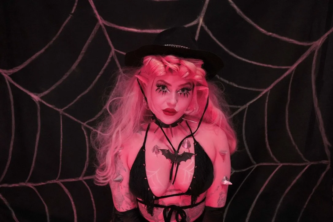 Woman with pink hair and makeup, dressed in a black costume with bat and spider web decorations, standing in front of a web-patterned background.