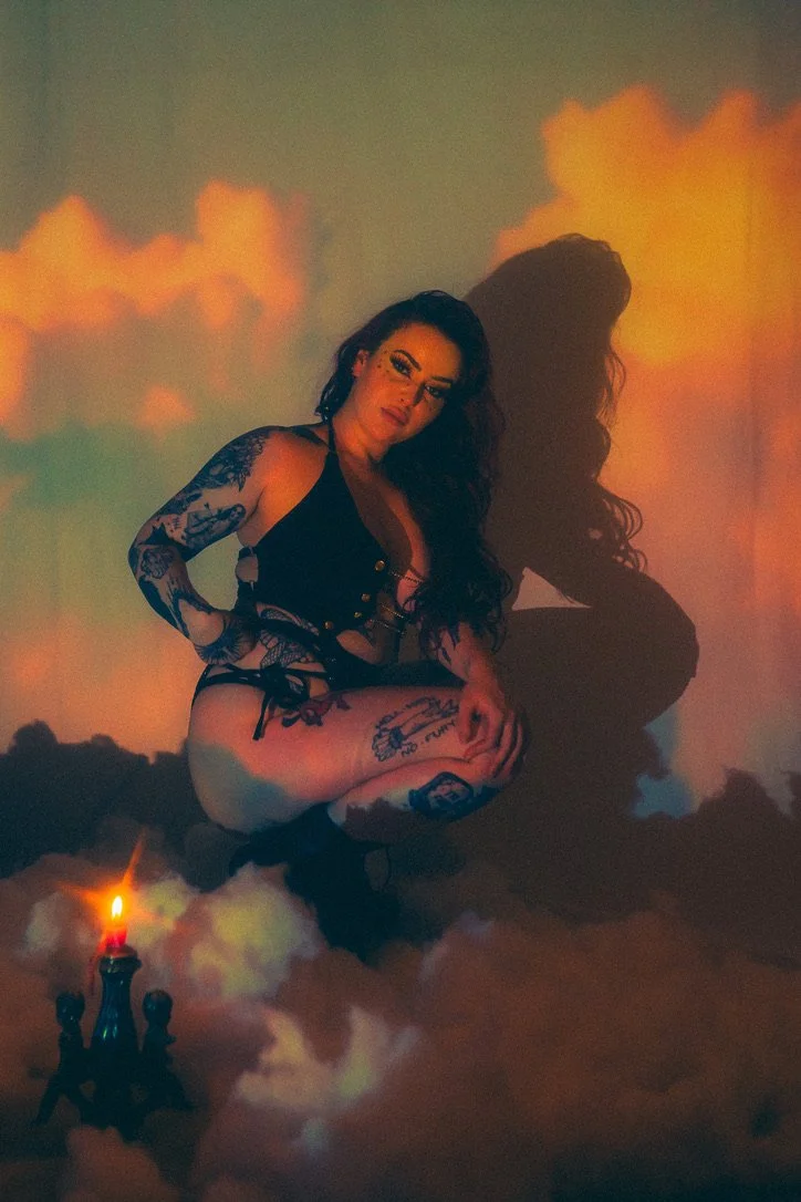 A woman with dark, curly hair and numerous tattoos, dressed in black lingerie, is kneeling on a cloud-like surface with a small, lit chandelier nearby. The background features colorful clouds, creating a dreamy, surreal atmosphere.