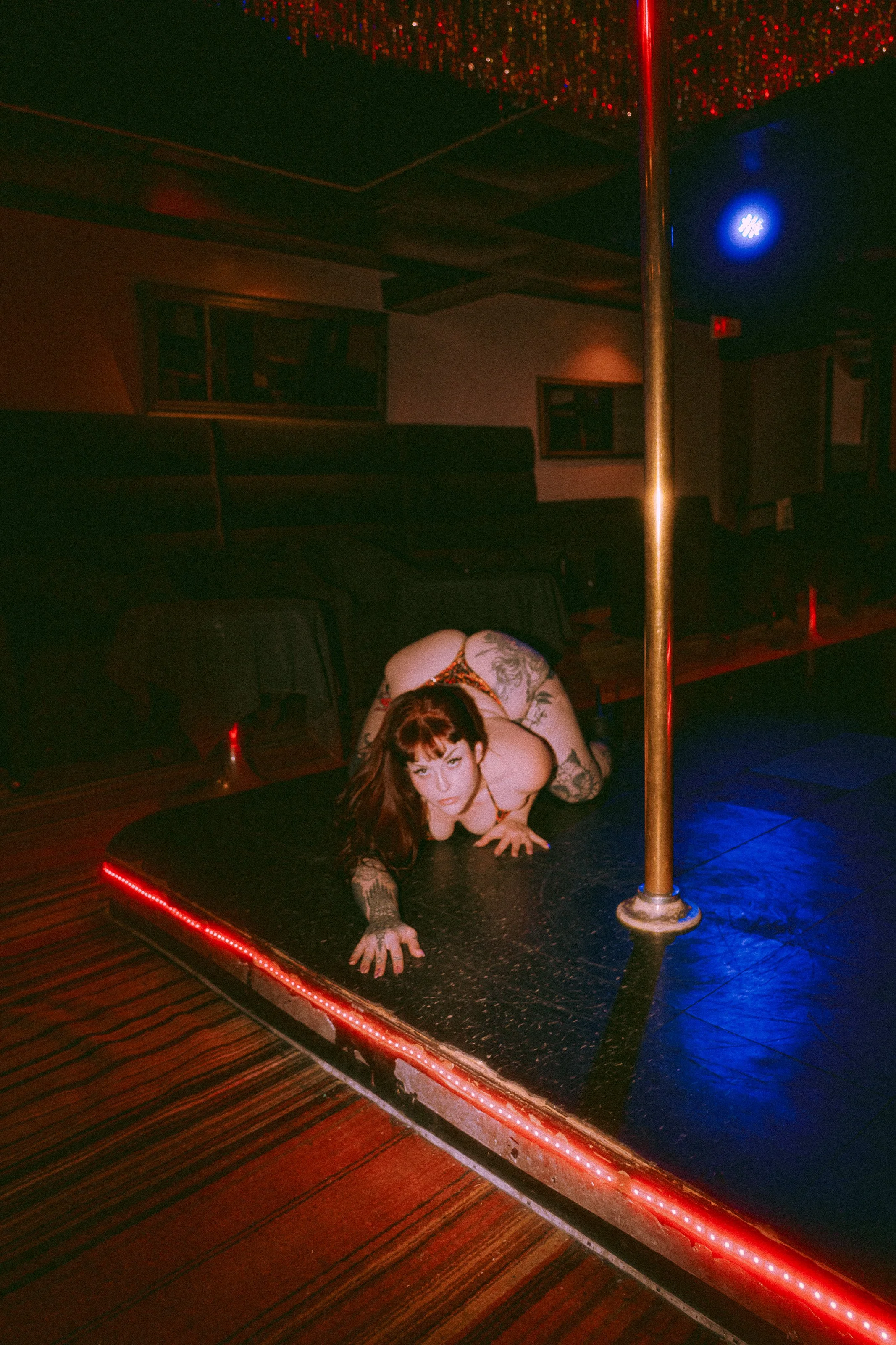 A woman with long hair and tattoos poses on a stage in a dimly lit club with a pole, red and blue neon lights, and seating in the background.