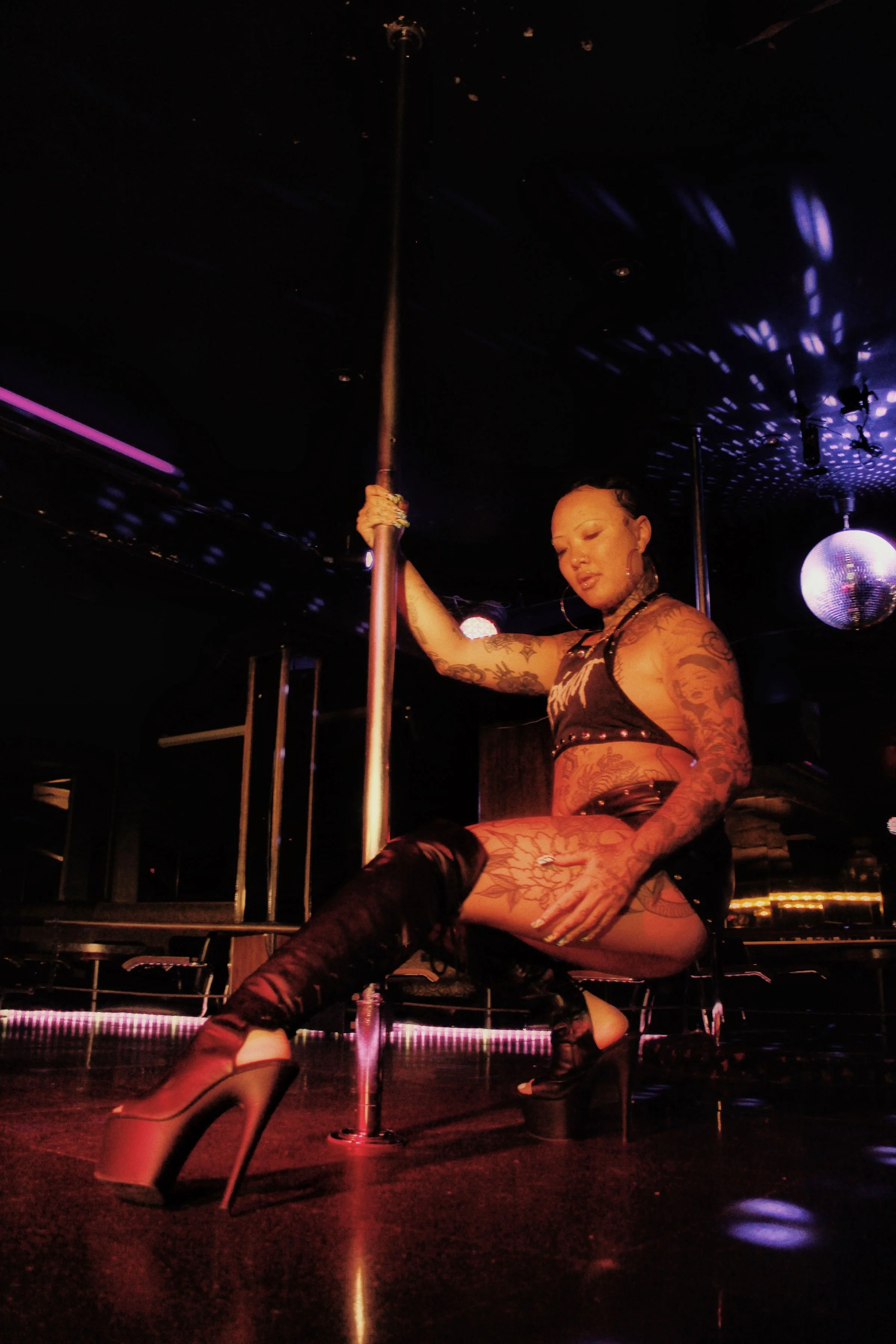 A performer with tattoos and high heels on a stage with a pole, in a nightclub setting with purple lighting and a disco ball.