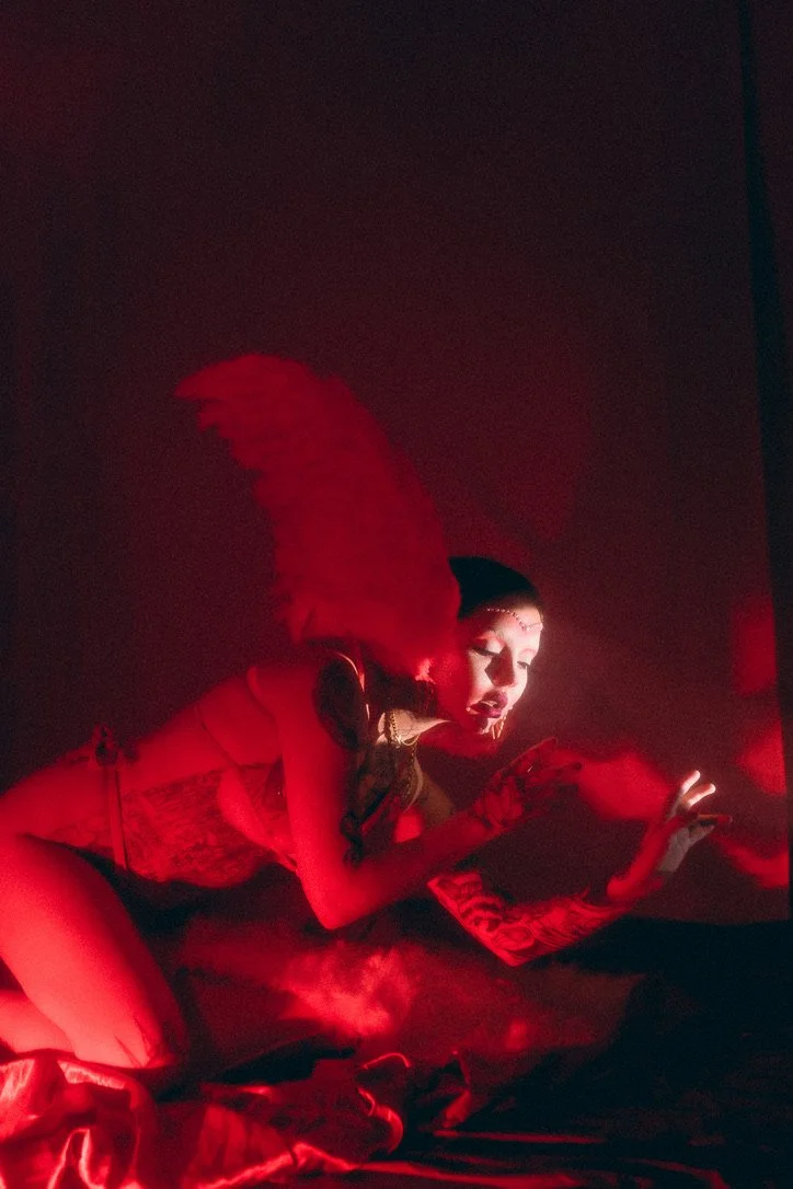 A performer dressed in a costume with a red feather headpiece, dark makeup, and jewelry, illuminated by red stage lighting.