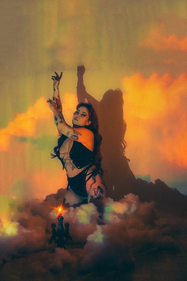 A woman with long dark hair and tattoos on her arms and side is posing with one arm raised. She is wearing a black outfit and is surrounded by clouds or smoke with a fiery glow. There is a candle or small flame in front of her, and her shadow is cast