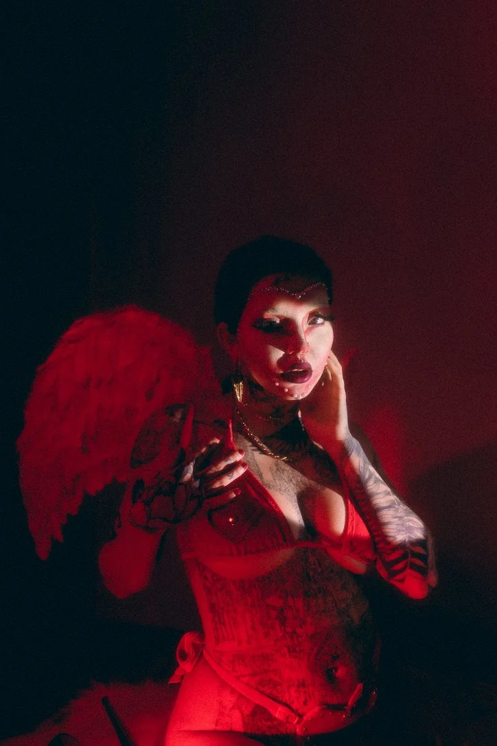 A woman with dark hair, tattoos, heavy makeup, and dark lipstick, wearing red lingerie and large red feathered wings, posing in a dimly lit red environment.