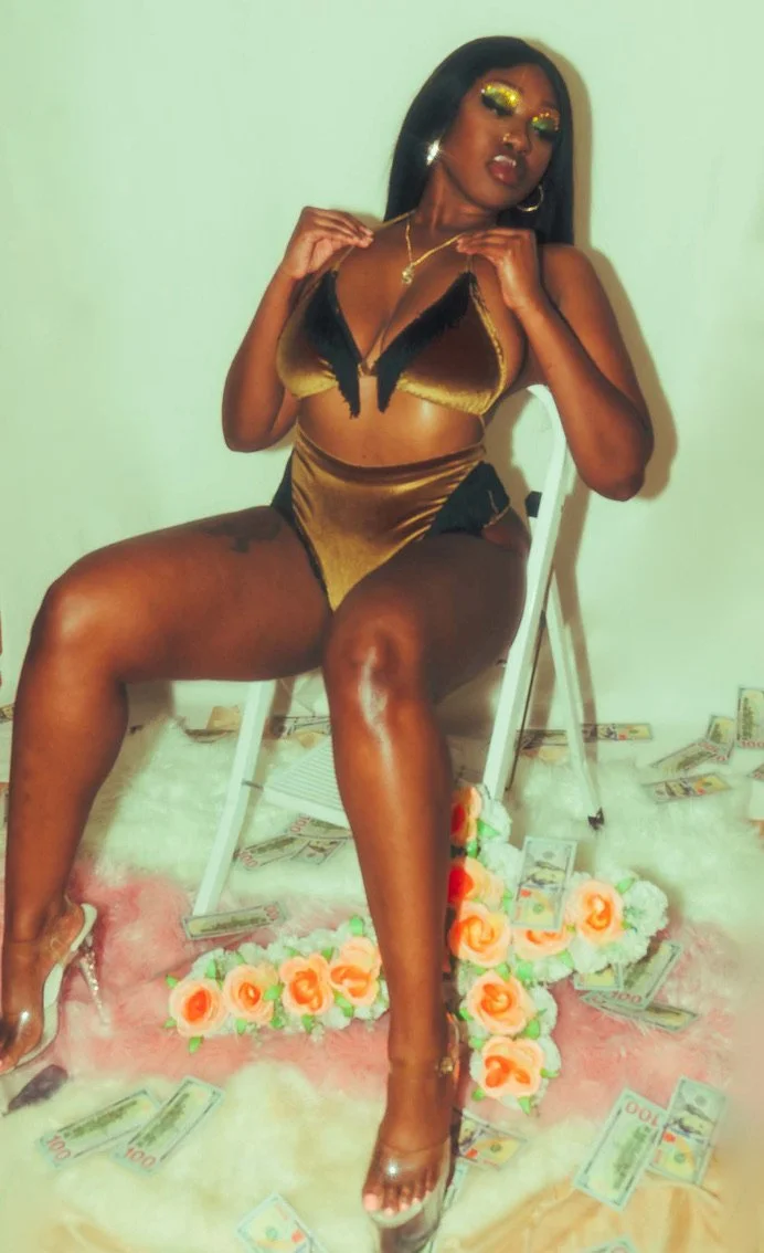 A woman dressed in gold and black lingerie sitting on a chair, surrounded by fake money and flowers on the floor.