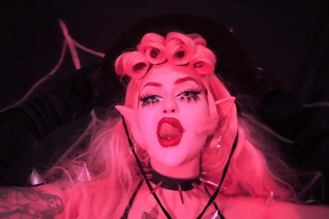 Person with pink hair styled in vintage curls, wearing a black hat and black clothing with spiked accessories, sticking out tongue with a piercing, and making a playful expression.