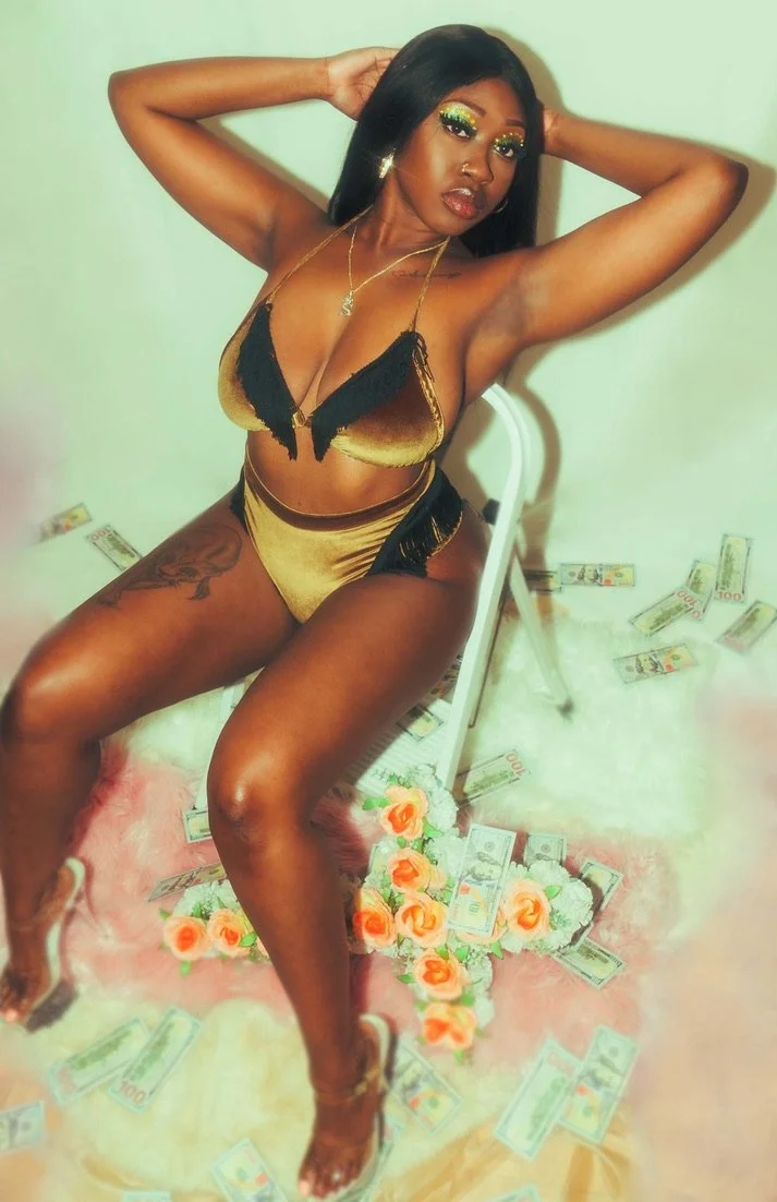 A woman in a gold and black lingerie set, sitting on a chair, surrounded by scattered US dollar bills and orange and white roses on a pink surface.