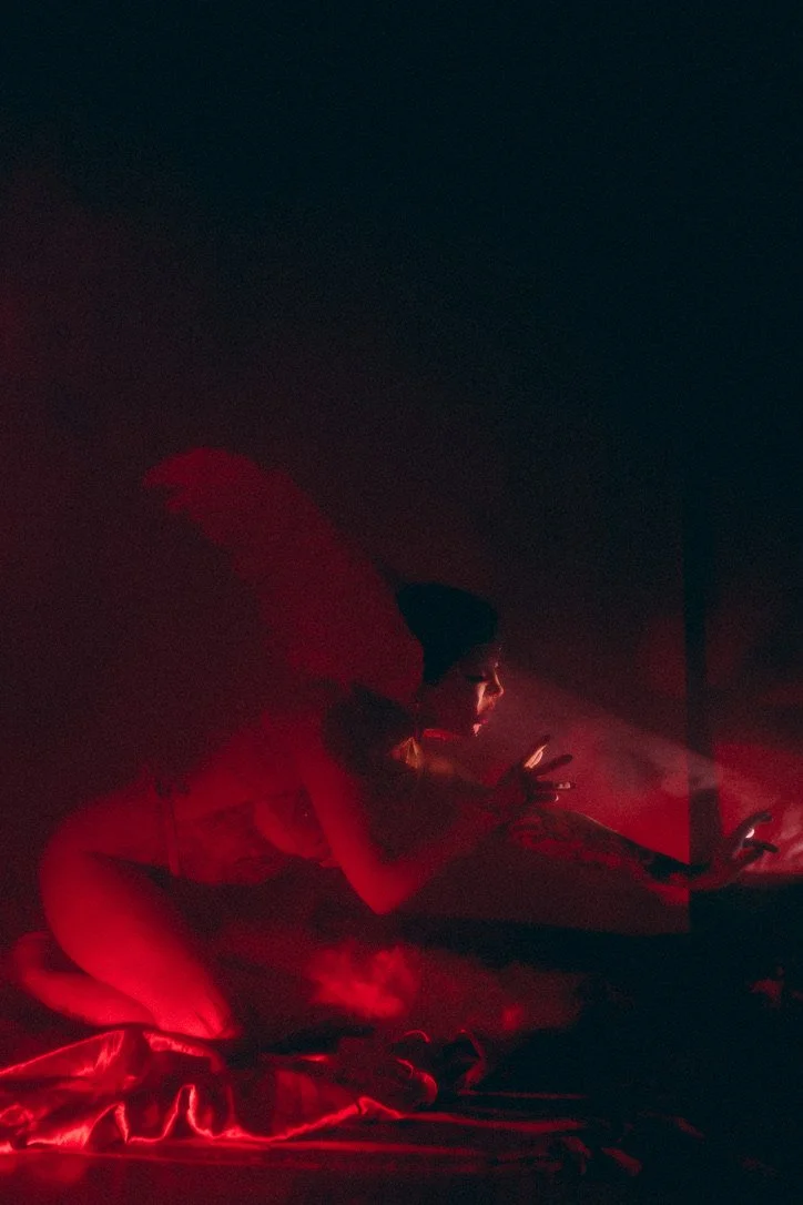 A person wearing a beanie and large red angel wings, illuminated by red neon lights, crouching and looking at a phone in a dark room.
