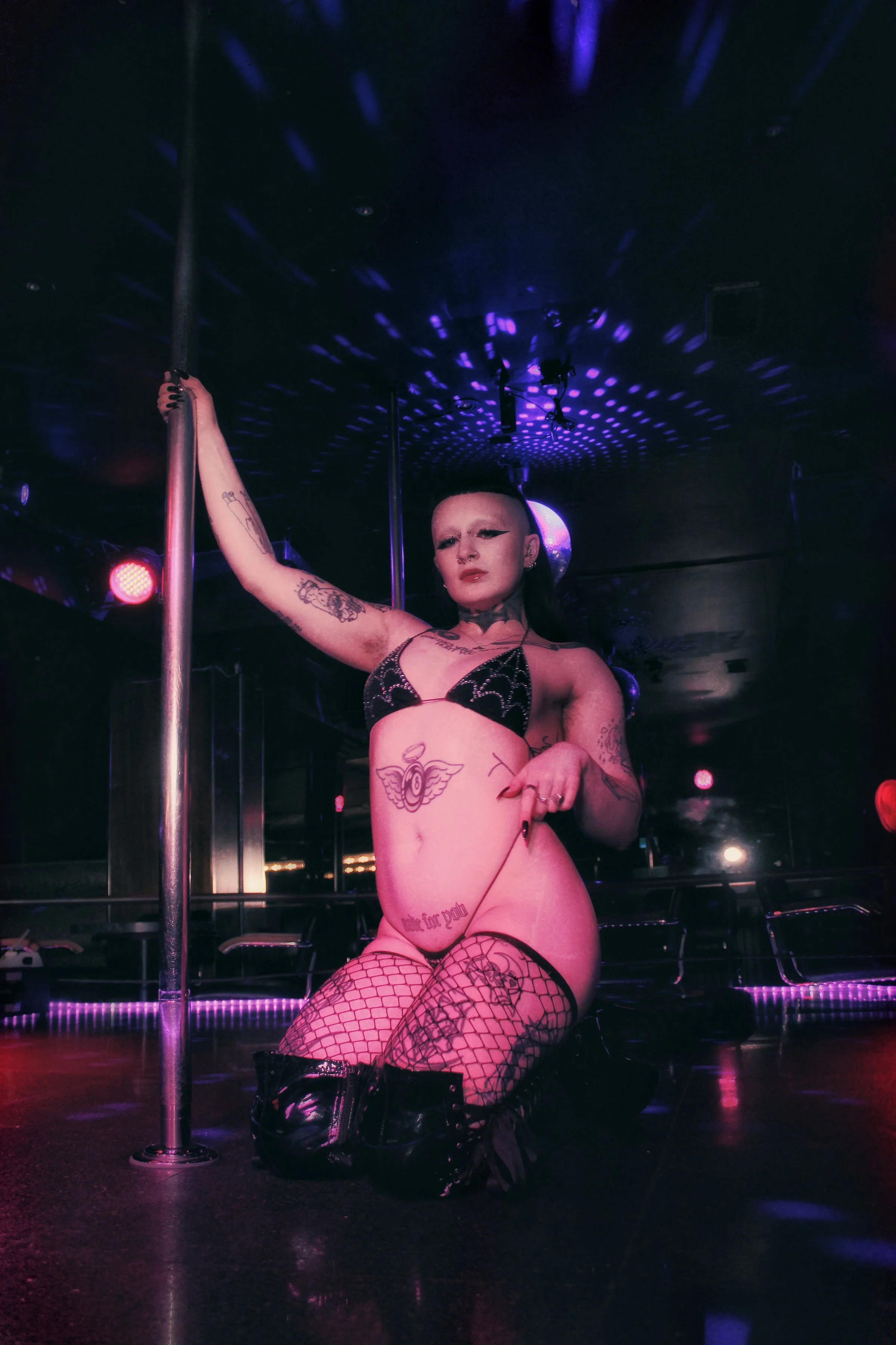 A woman with tattoos and fishnet stockings performing a dance on a stage with a pole in a nightclub, illuminated by colorful lights.