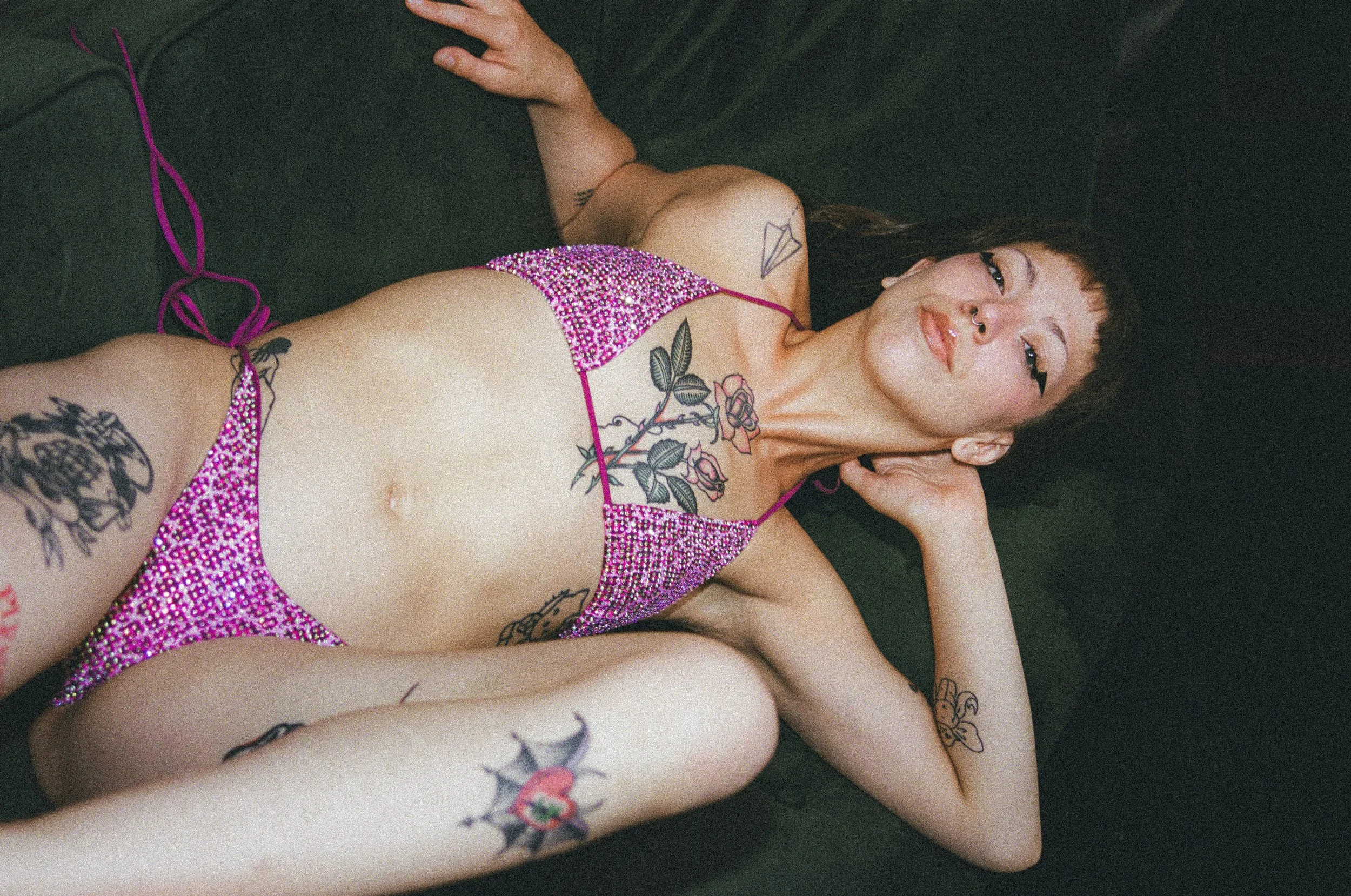 Tattooed person reclining in a sparkling pink outfit against a black background
