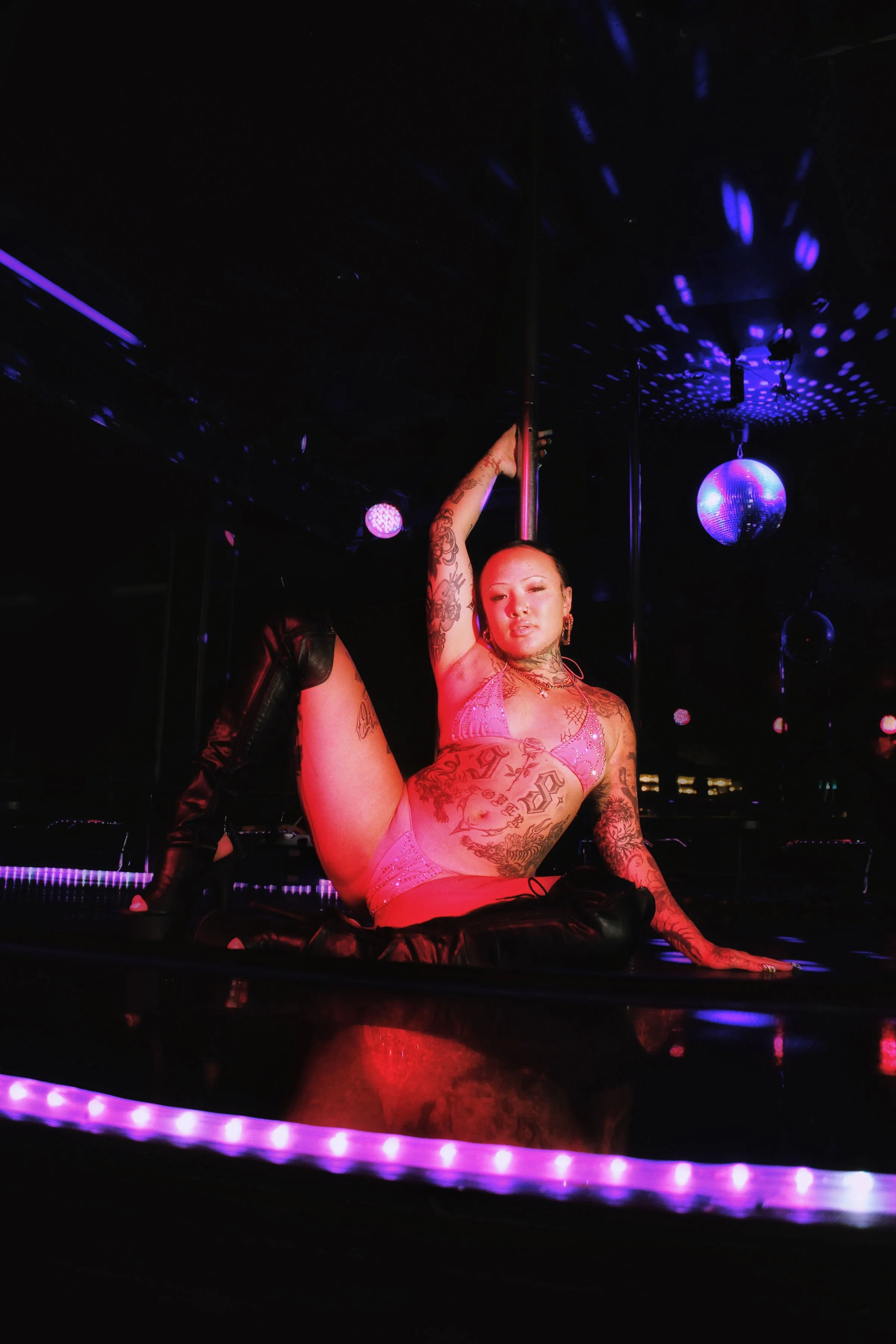 A woman with tattoos on her arms and body, wearing a pink lingerie set, is posing on the floor of a dimly lit nightclub or dance studio with a pole, stage lighting, and a disco ball overhead.