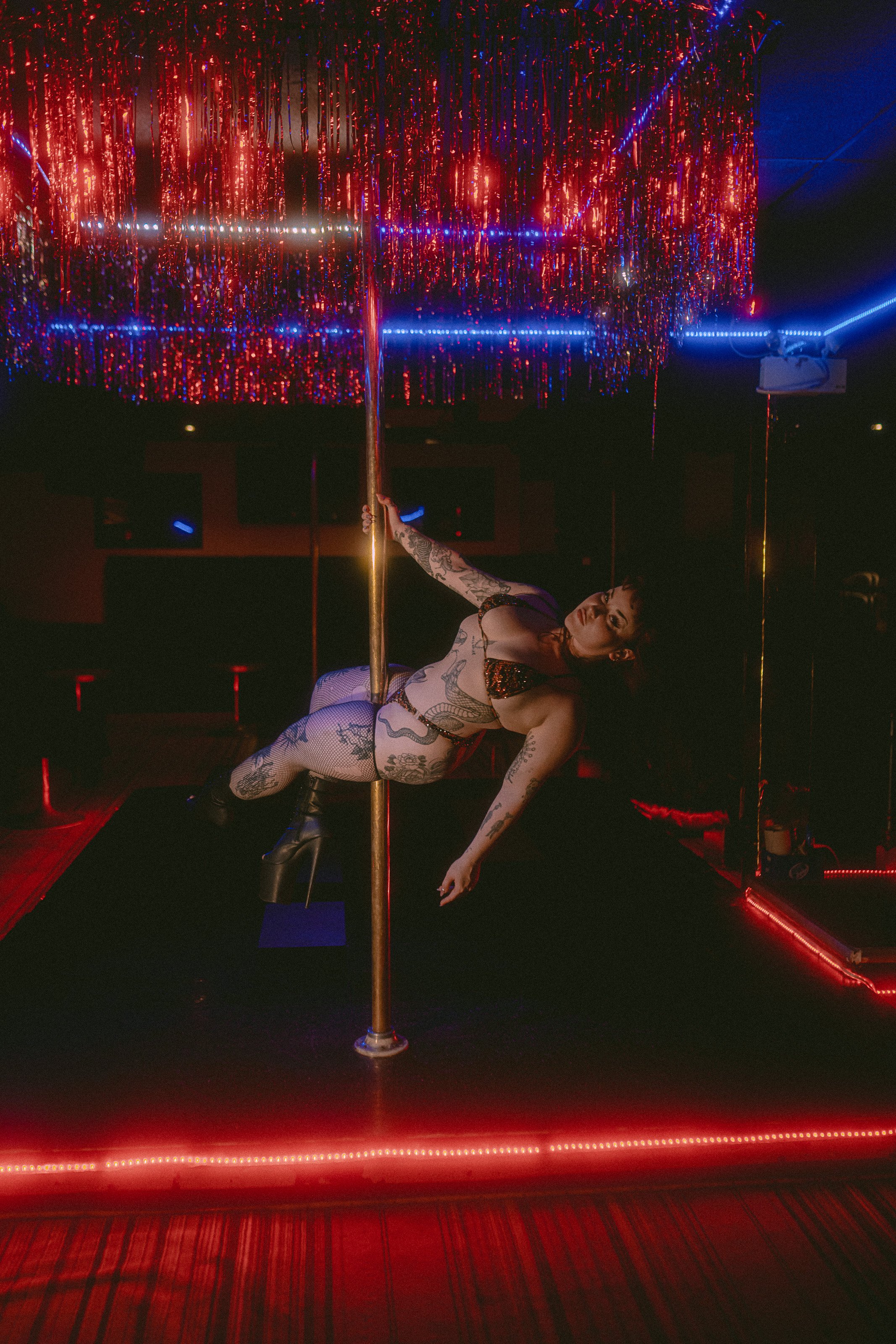 A woman with tattoos and fishnet stockings performs a pole dance under red and blue neon lights in a dimly lit club or entertainment venue.